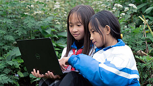 Zwei Mädchen aus Vietnam im blauen Schuloutfit lernen zusammen draußen am Laptop im Grünen. Zwei Mädchen aus Vietnam sitzen in der Natur und lernen gemeinsam am Laptop, umgeben von grünen Pflanzen und Blumen.