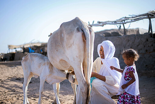 Eine Frau sitzt auf einem Melkschemel neben einer Kuh und einem Kalb. Neben ihr steht ihre Tochter.