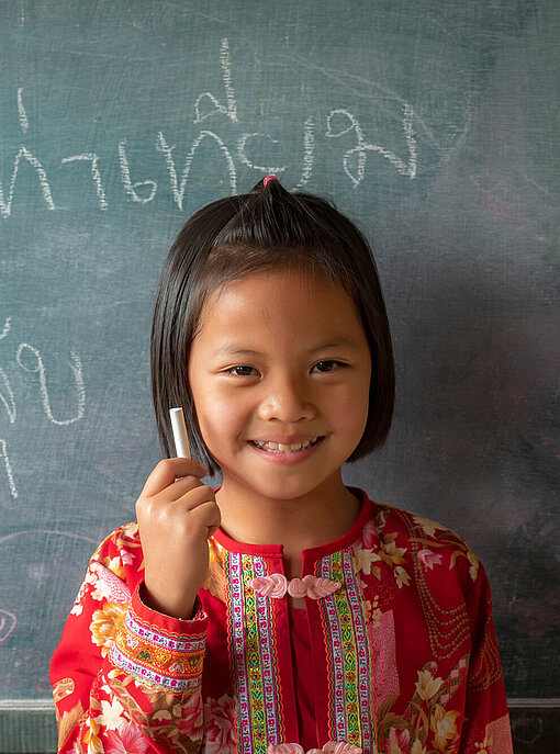 Die Schulen in Chiang Rai sind inzwischen besser gegen Naturkatastrophen gewappnet. Ein Mädchen steht grinsend vor einer Tafel. Sie hat ein Stück Kreide in der Hand.