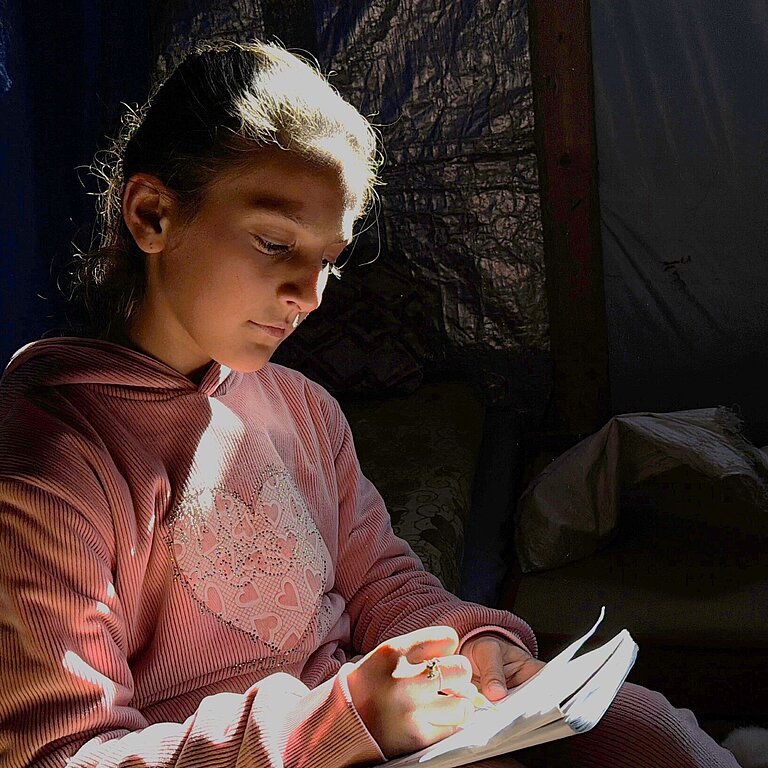 Ein Mädchen sitzt in einem dunklen Zelt und malt ein Bild, ein einzelner Lichtstrahl fällt auf ihr Gesicht