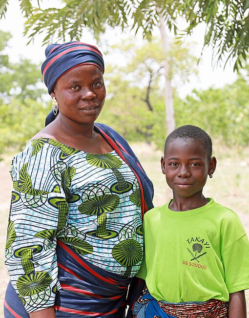 Eine Frau in bunten Kleider steht mit ihrer Tochter in einem T-Shirt unter einem Baum
