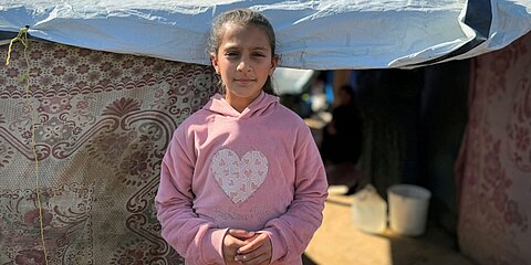 Ein Mädchen steht vor einem provisorischen Zelt, das mit einer Plane und dekorativen Stoffen abgedeckt ist. Das Kind trägt einen rosa Kapuzenpullover mit einem großen Herz auf der Brust. Im Hintergrund sieht man einen sandigen Boden, einen weißen Eimer und im Halbschatten weitere Personen sowie den Eingang zum Zelt.