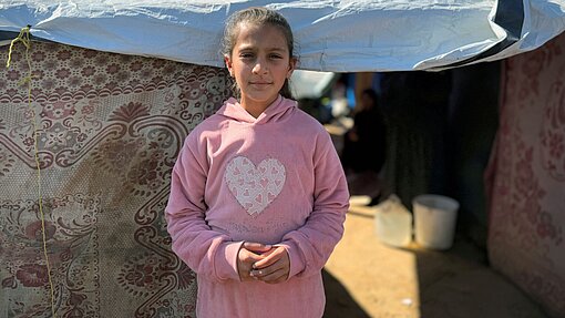 Ein Mädchen steht vor einem provisorischen Zelt, das mit einer Plane und dekorativen Stoffen abgedeckt ist. Das Kind trägt einen rosa Kapuzenpullover mit einem großen Herz auf der Brust. Im Hintergrund sieht man einen sandigen Boden, einen weißen Eimer und im Halbschatten weitere Personen sowie den Eingang zum Zelt.