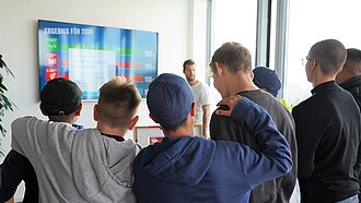 Eine Gruppe Schüler steht im Halbkreis vor einem großen Bildschirm. Man sieht die Schüler von hinten.