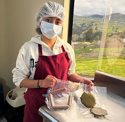 Eine junge Frau trägt eine Schürze, Mund- und Haarschutz und hält pflanzliche Würtchen und Burger Pattys hoch.
