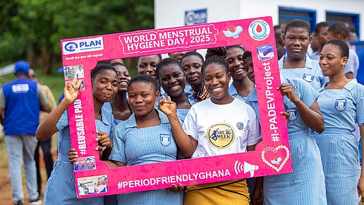 Mehrere Mädchen in Schuluniform stehen beieinander und lachen durch einen pinken Rahmen in die Kamera. Der Rahmen macht auf den World Menstrual Hygiene Day aufmerksam.