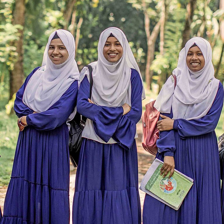 Mädchen mit blauen Kleidern und weißen Kopftüchern lachen in die Kamera und halten Schulbücher in den Händen