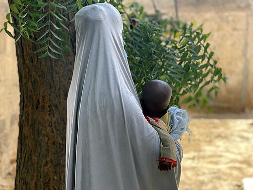 Eine Frau ist von hinten zu sehen, sie trägt ein Kopftuch und ist nicht zu erkennen. Sie hält Baby im Arm.