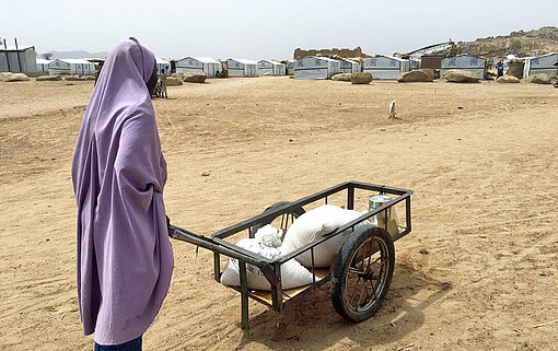Eine Frau im weißen Baumwollkleid schiebt einen Holzkarren über einen sandigen Weg