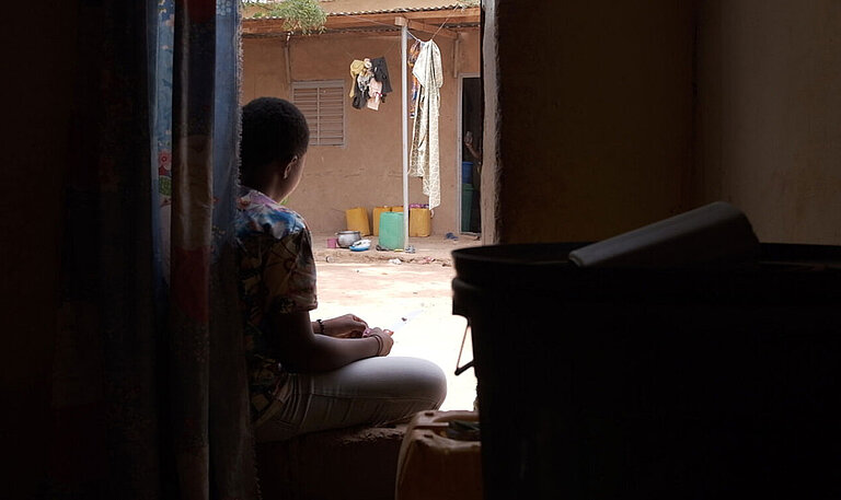 Durch einen Hauseingang sieht man eine junge Frau vor der Tür im Hof sitzen.