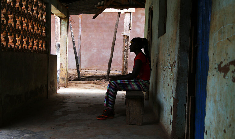 Eine Frau sitzt vor einem Haus unter einem Vordach, man sieht nur ihre schwarze Silhouette 