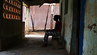 Eine Frau sitzt vor einem Haus unter einem Vordach, man sieht nur ihre schwarze Silhouette 