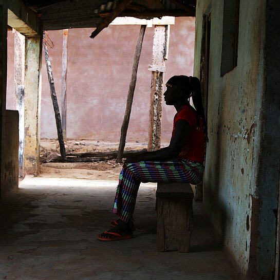 Eine Frau sitzt vor einem Haus unter einem Vordach, man sieht nur ihre schwarze Silhouette 