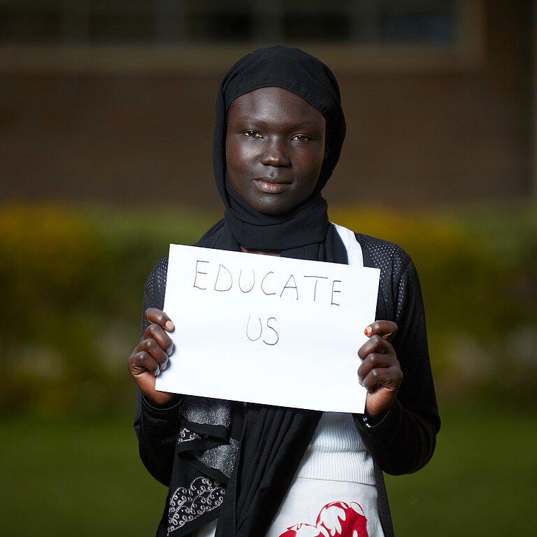 Ein Mädchen hält ein Schild hoch, auf dem Educate Us (bildet uns) steht