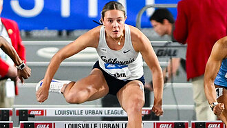 Die Sportlerin Maria Testa im Trikot mit der Aufschrift „California“ überspringt fokussiert eine Hürde während eines Wettkampfs. Im Hintergrund sind weitere Läufer:innen und Offizielle zu sehen.