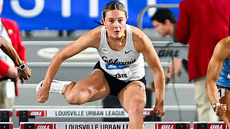 Die Sportlerin Maria Testa im Trikot mit der Aufschrift „California“ überspringt fokussiert eine Hürde während eines Wettkampfs. Im Hintergrund sind weitere Läufer:innen und Offizielle zu sehen.