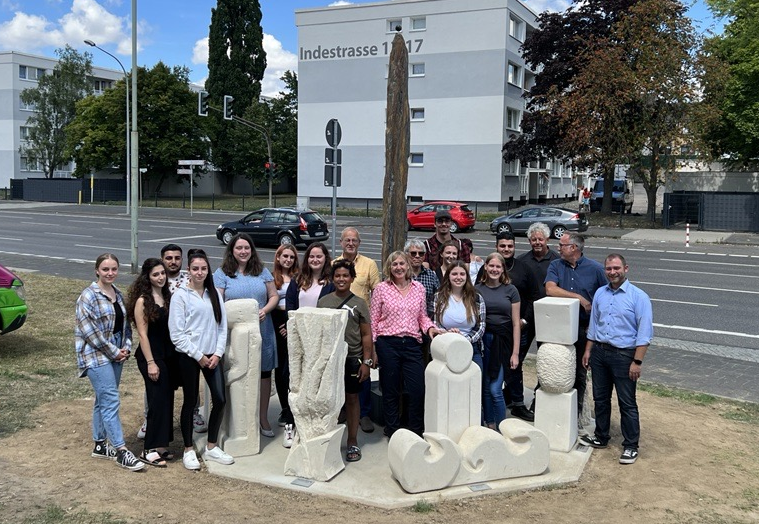 Eine Gruppe Menschen steht am Mahnmal zur Flutkatastrophe in Eschweiler. Es besteht aus einem Monolithen, der von kleineren Skulpturen umrundet ist.