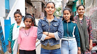 Fünf jugendliche Mädchen aus Indien stehen in einer engen Straße mit heruntergekommenen Gebäuden. Sie haben jeweils eine Hand auf die Schulter der anderen gelegt. Die Jugendliche in der Mitte hat ihre Arme verschränkt, alle schauen entschlossen.