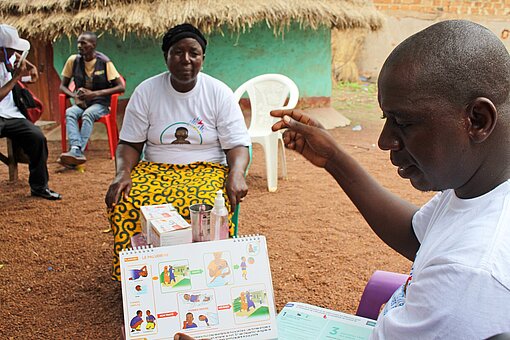 Aufklärung über Malaria Ein Mann hält ein Faktenblatt mit Symbolen in der Hand und erklärt einer Frau, die ihm gegenüber sitzt, etwas über Malaria