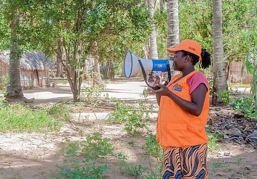 Eine mosambikanische Frau in einer orangenen Warnweste spricht in ein Megaphon