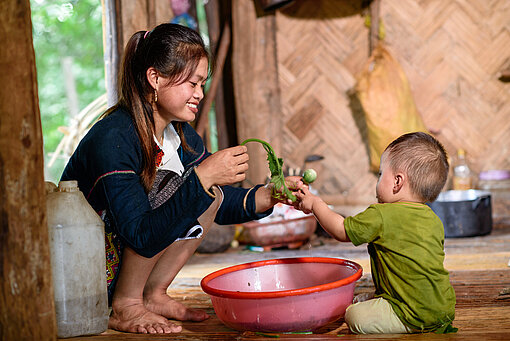 eine Frau sitzt in der Hocke vor einer Schüssel mit Wasser und putzt darin Gemüse, ein Kleinkind sitzt neben der Schüssel und macht mit