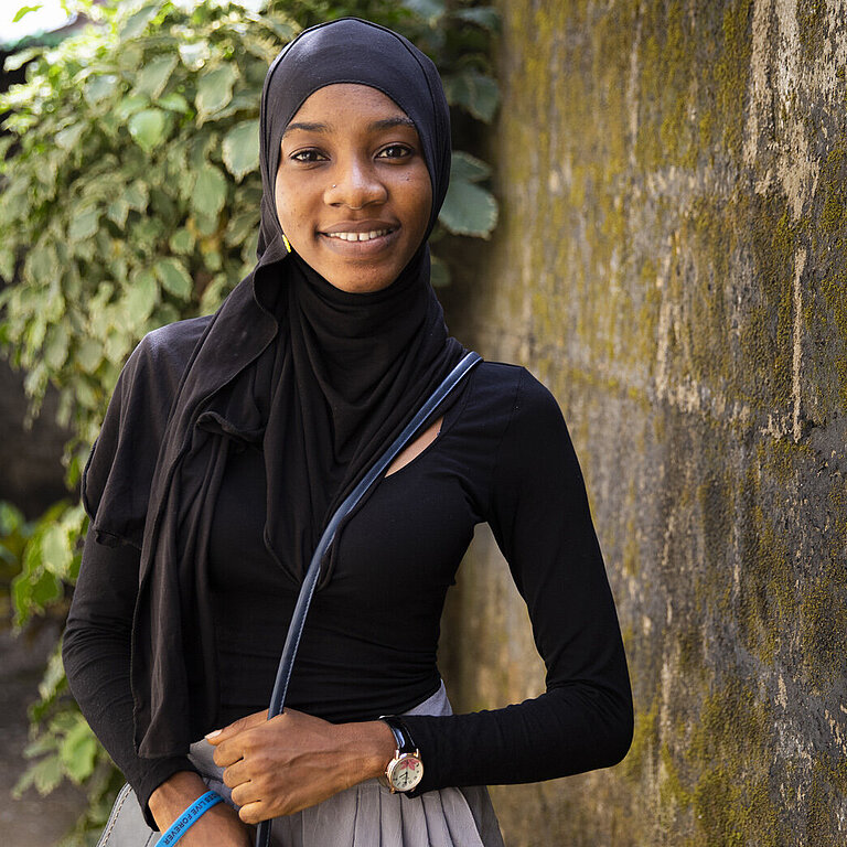Portrait von Isatu vor einer Mauer und Pflanze.