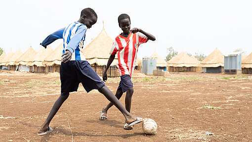 Akim und Lim haben sich im Flüchtlingslager kennengelernt und verbringen viel Zeit zusammen an den kinderfreundlichen Orten. Lim und Akim spielen mit einem Fußball.