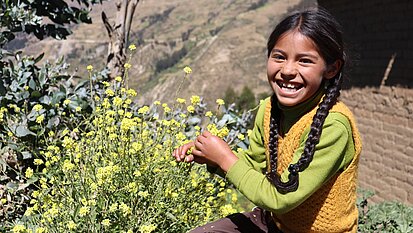Ein Mädchen sitzt vor einer Berglandschaft in Peru. Sie lacht freudig und hält den Stiel einer gelben Blume in der Hand.