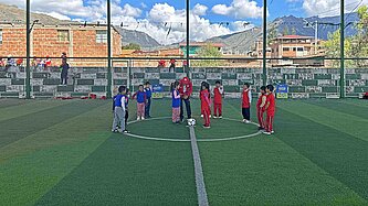 Mädchen und Jungen stehen in Trikots auf einem überdachten Fußball-Platz, im Hintergrund sind schneebedeckte Berge sichtbar.