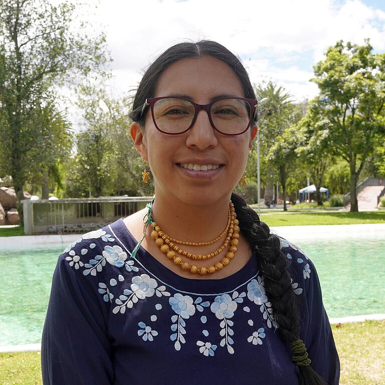 Verónica steht im Freien vor einer Wasserquelle in einer ländlichen Umgebung und blickt in die Kamera