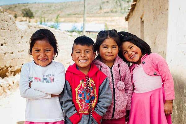 Drei Mädchen und ein Junge stehen vor ihrem Haus in Bolivien. Sie stehen eng beieinander und lachen.