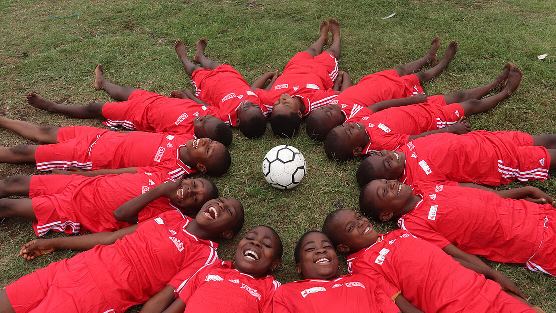 Kinder in roten Trikots liegen im Kreis um einen Fußball herum. Ihre Köpfe zeigen in die Mitte des Kreises, ihre Füße nach außen.