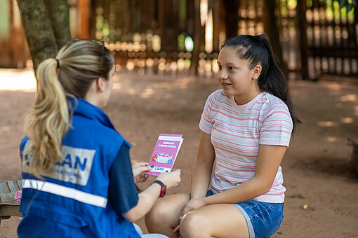 Ein Mädchen sitzt einer Frau gegenüber, welche eine Plan-Weste trägt und einen Flyer über Menstruationshygiene in der Hand hält.