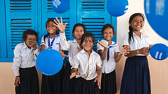 Bild: Mehrere Schülerinnen aus Kambodscha stehen vor ihrer Schule und werfen Luftballons in Richtung Kamera