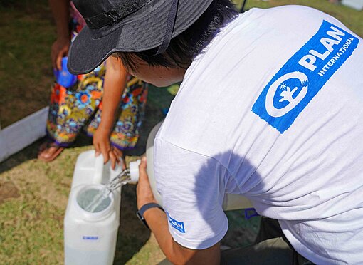 Eine Frau mit dem blauen Logo von Plan International auf dem T-Shirt gießt frisches Trinkwasser in einen Kanister