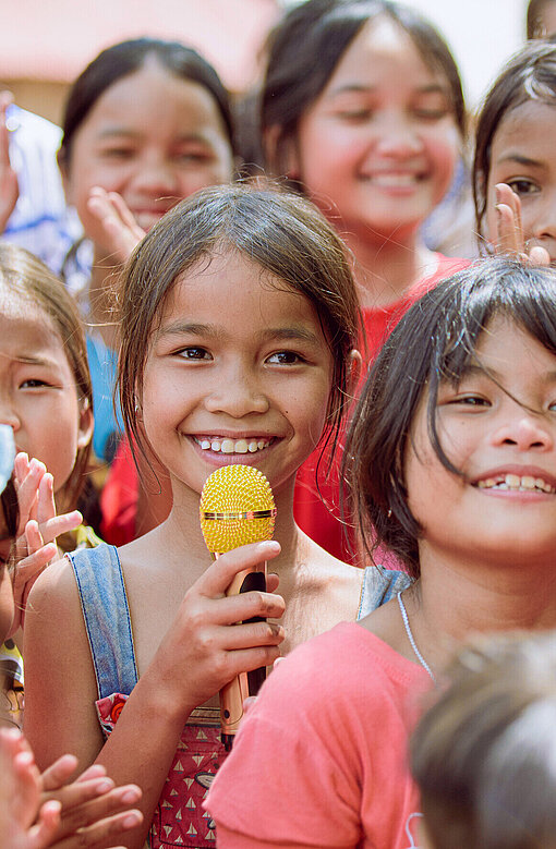 Mädchen in einer Gruppe, eine Teilnehmerin mit Mikrofon, lernen im Kinder Club in Vietnam praktische und soziale Fähigkeiten, unterstützt durch Plan International.