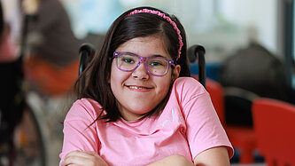 Ein Mädchen sitzt in einem Rollstuhl, es lächelt in die Kamera und hat die Hände vor dem Körper verschränkt