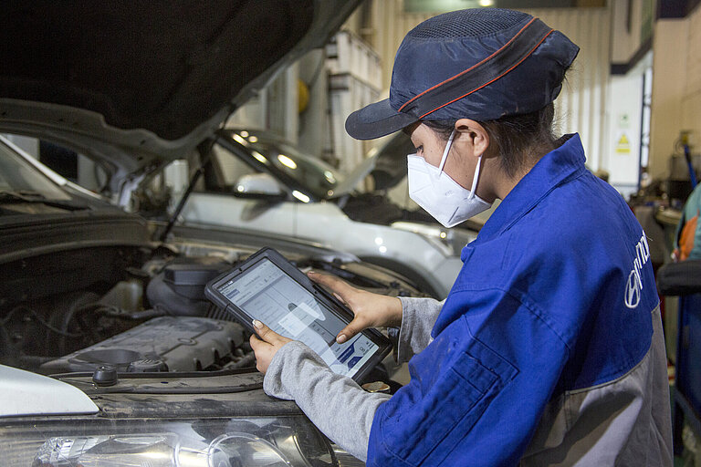 Eine Frau mit Kappe, FFP-2-Maske und in Arbeitskleidung steht vor einer geöffneten Motorhaube und trägt etwas in ein Tablet ein