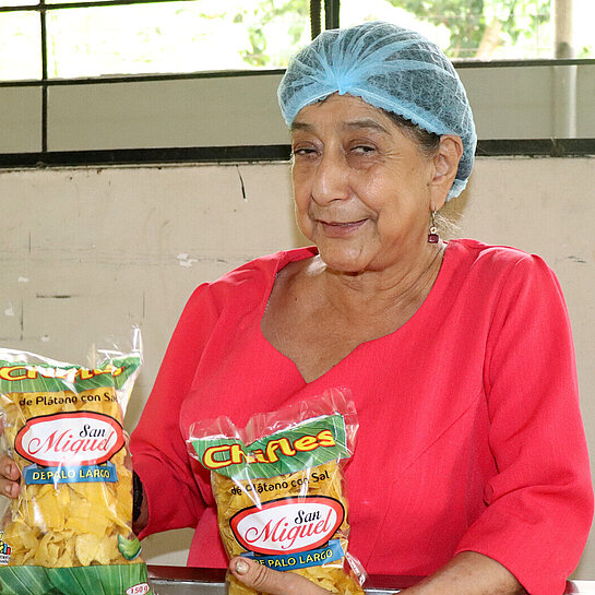 Eine Frau in einer bunten Bluse hält zwei Tüten mit Bananenchips in den Händen. Sie trägt ein Haarnetz.