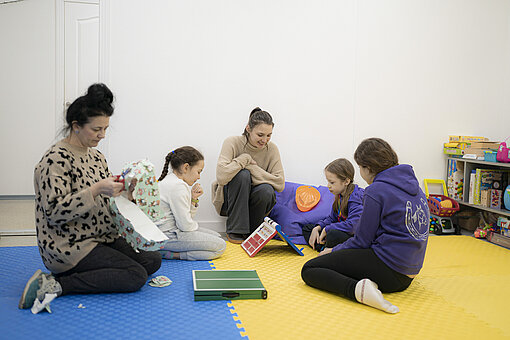 Zwei Frauen sitzen mit drei Kindern in einem hellen Raum auf buntem Boden und spielen