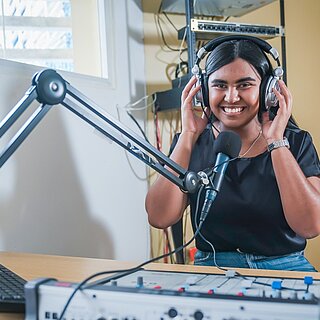 Edith sitzt an einem Radio-Mischpult, spricht in ein Mikrofon und moderiert eine Sendung für Jugendliche. Alternative Bildunterschrift: