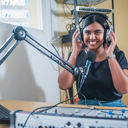 Edith sitzt an einem Radio-Mischpult, spricht in ein Mikrofon und moderiert eine Sendung für Jugendliche. Alternative Bildunterschrift: