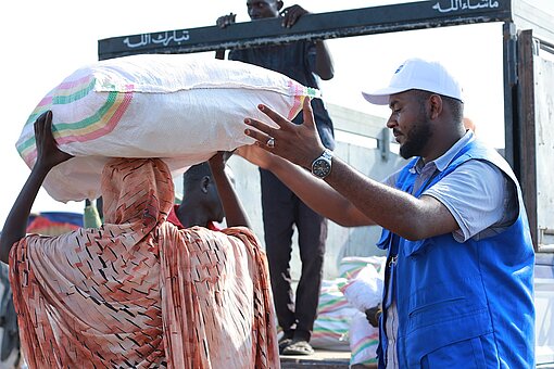 Ein Mitarbeiter von Plan International Sudan reicht einer Frau einen großen Sack Hilfslieferung, den sie auf dem Kopf trägt