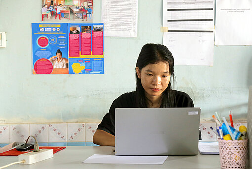 Kann sitzt an einem Schreibtisch, vor ihr ein Laptop - Sie schaut konzentriert auf dessen Bildschirm. 