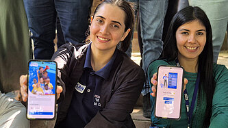 Zwei Frauen sitzen nebeneinander und halten jeweils ihre Handys in die Kamera. 