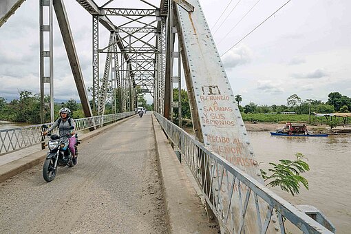Eine einspurige Straßenbrücke mit einem Stahlbogen überspannt einen Fluss mit bräunlichem Wasser.