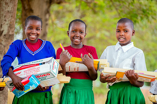 Drei Mädchen mit Schuluniformen halten Hefte, Stifte, Seife und Menstruationsprodukte in den Armen.
