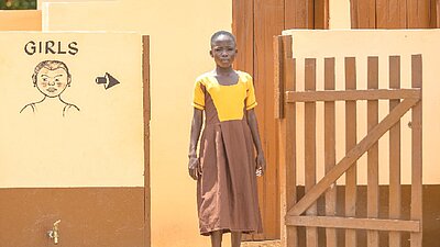 Ein Mädchen steht vor einem gelben Toilettenhäuschen mit braunen Türen. Links ist ein Schild mit einer Zeichnung und dem Wort „GIRLS“ angebracht, das auf die Mädchentoiletten hinweist.