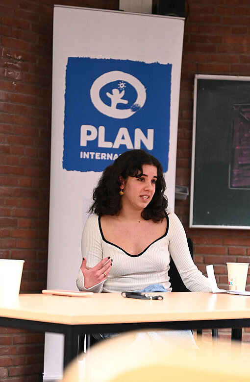 Lale Ahmerkamp ruft die Teilnehmer:innen zur Solidarität mit den Protestierenden im Iran auf. Eine junge Frau sitzt hinter einem Tisch und spricht zum Publikum.