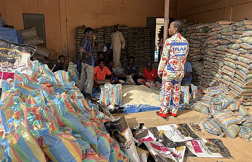 Ein sudanesischer Mann mit Plan-Logo auf der Jacke steht zwischen Bergen von Hilfslieferungen